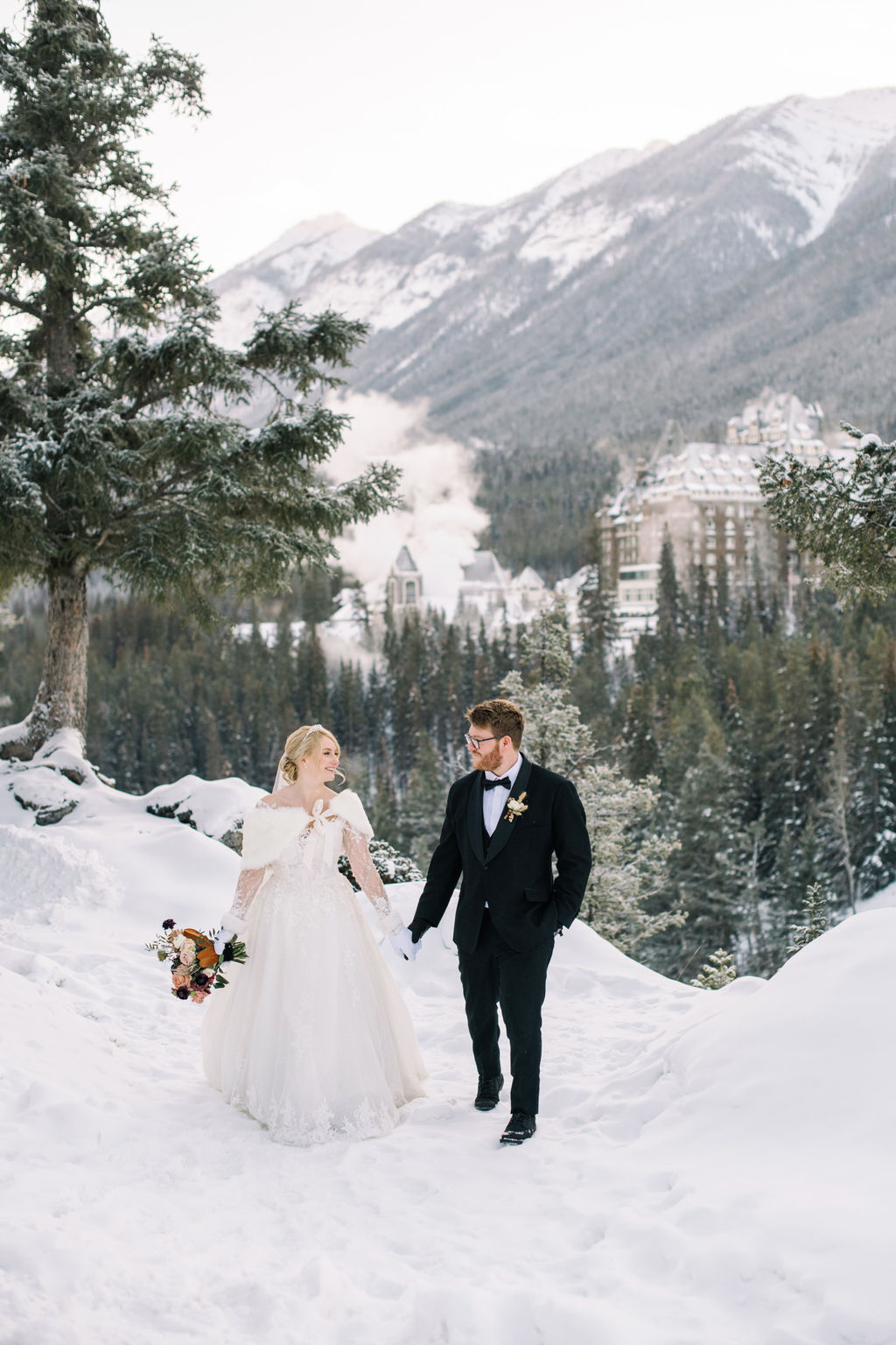 Bride and groom pose in the snow-dusted mountains with Fairmont Banff Springs in the distance, capturing the magic of a winter wedding in Banff.