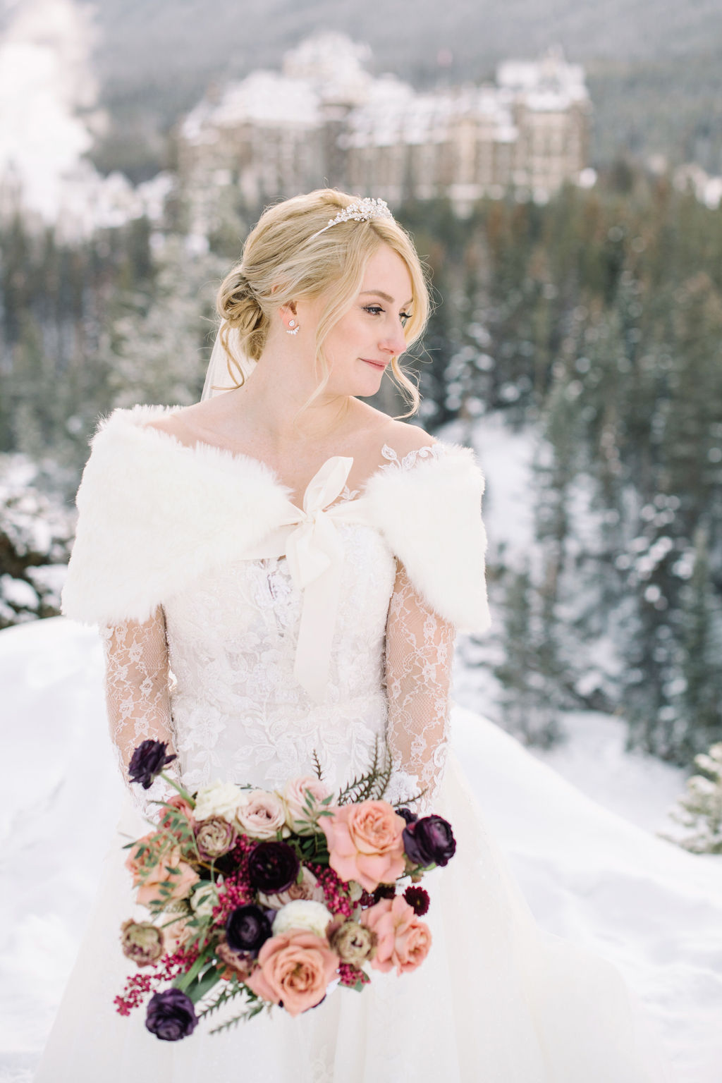 Bride poses with her blush and burgundy bouquet in the snow-dusted mountains with Fairmont Banff Springs in the distance.
