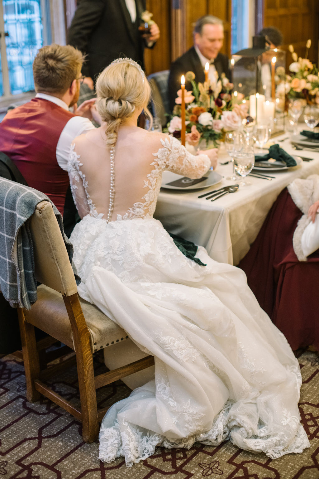The newlyweds enjoy dinner with their guests at a long candlelit table surrounded by warm tones and festive details. Cozy winter reception in the Angus Room, styled with rich hues and elegant decor for an intimate wedding celebration.
