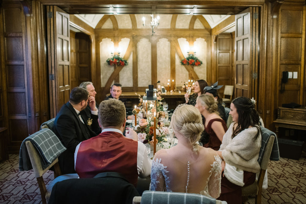 The newlyweds enjoy dinner with their guests at a long candlelit table surrounded by warm tones and festive details. Cozy winter reception in the Angus Room, styled with rich hues and elegant decor for an intimate wedding celebration.