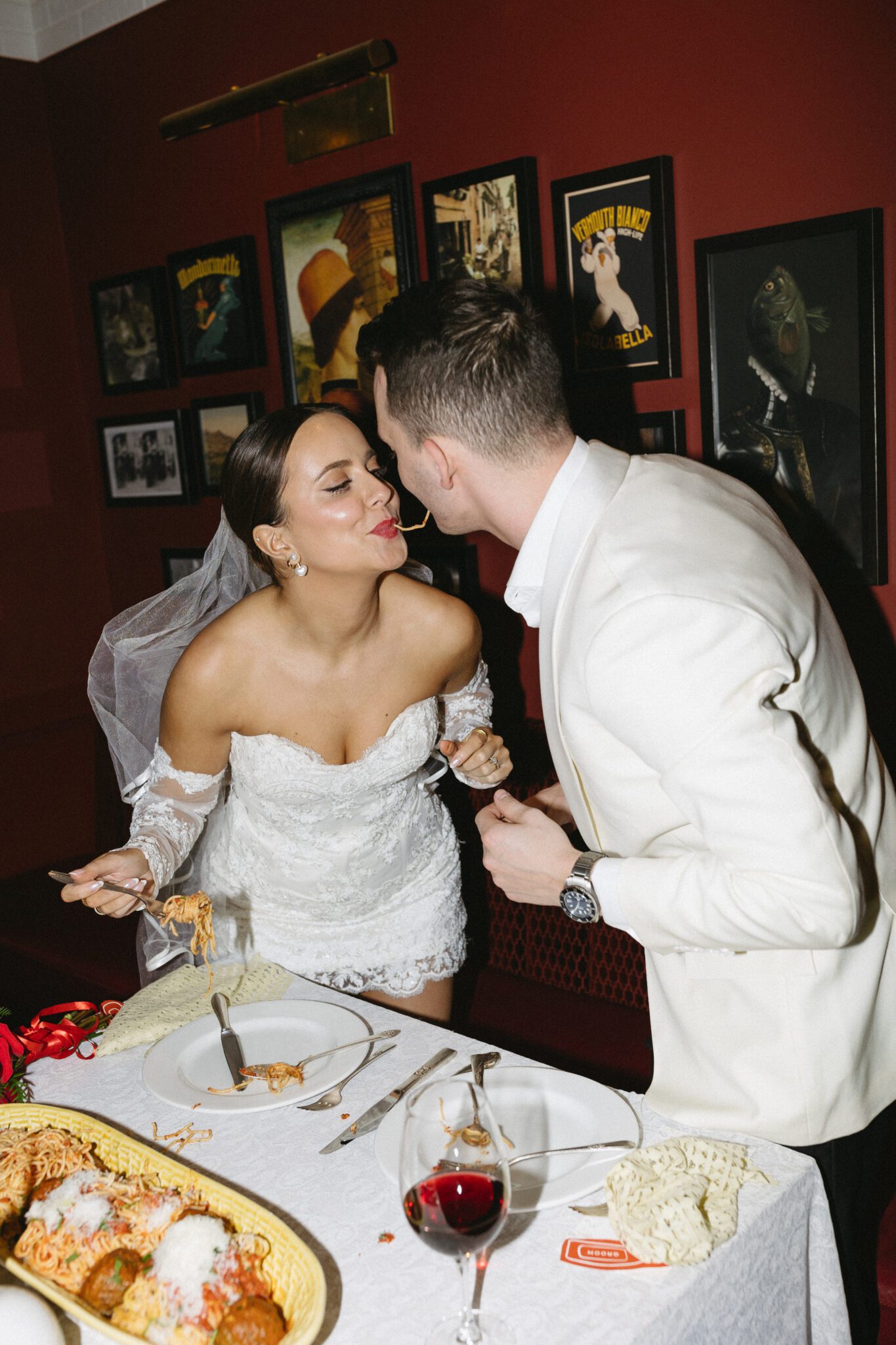 This Italy inspired wedding inspiration at Buffo Restaurant in Calgary, showcases modern sophistication, vintage charm, and a vibrant red colour palette. This photo shows the bride and groom eating spaghetti at the reception. #brideandgroomattire #modernweddinginspiration #receptiontoast #restaurantreception #vintageweddinginspo