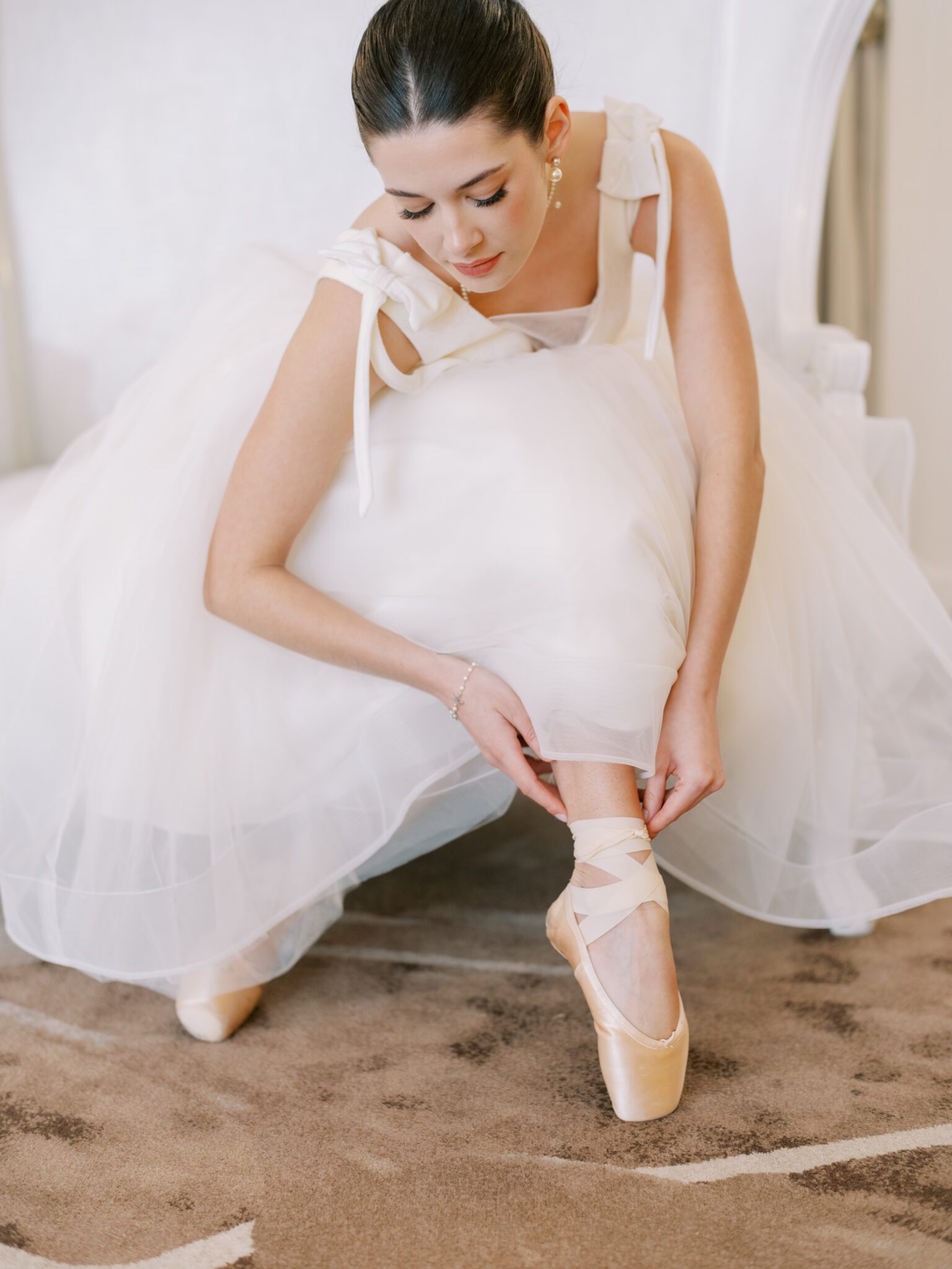 A ballerina in a white tutu sits on an elegant white settee, gracefully adjusting her pink pointe shoe. She has her dark hair in a bun and is surrounded by light-colored, sophisticated decor.