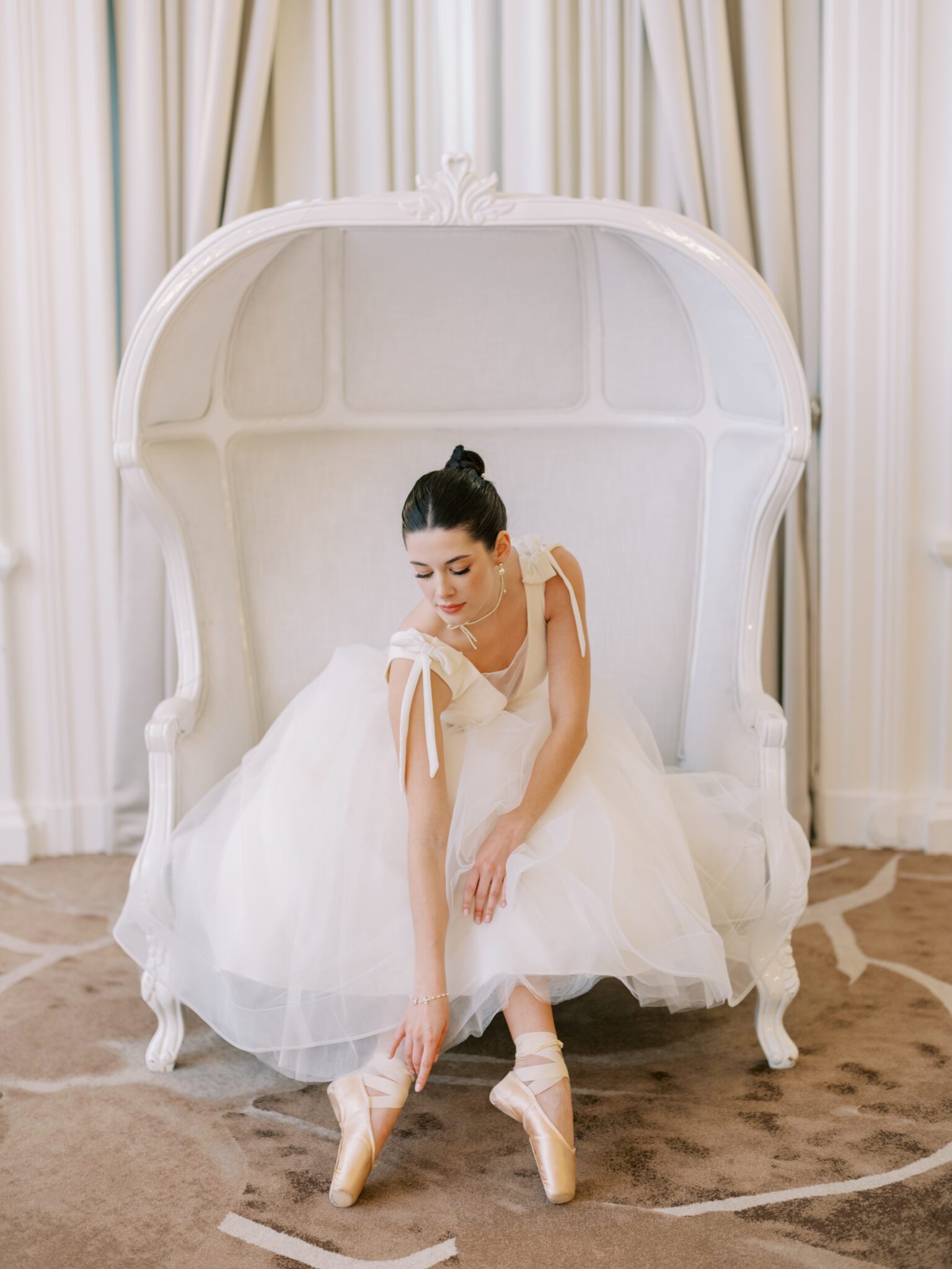 A ballerina in a white tutu sits on an elegant white settee, gracefully adjusting her pink pointe shoe. She has her dark hair in a bun and is surrounded by light-colored, sophisticated decor.