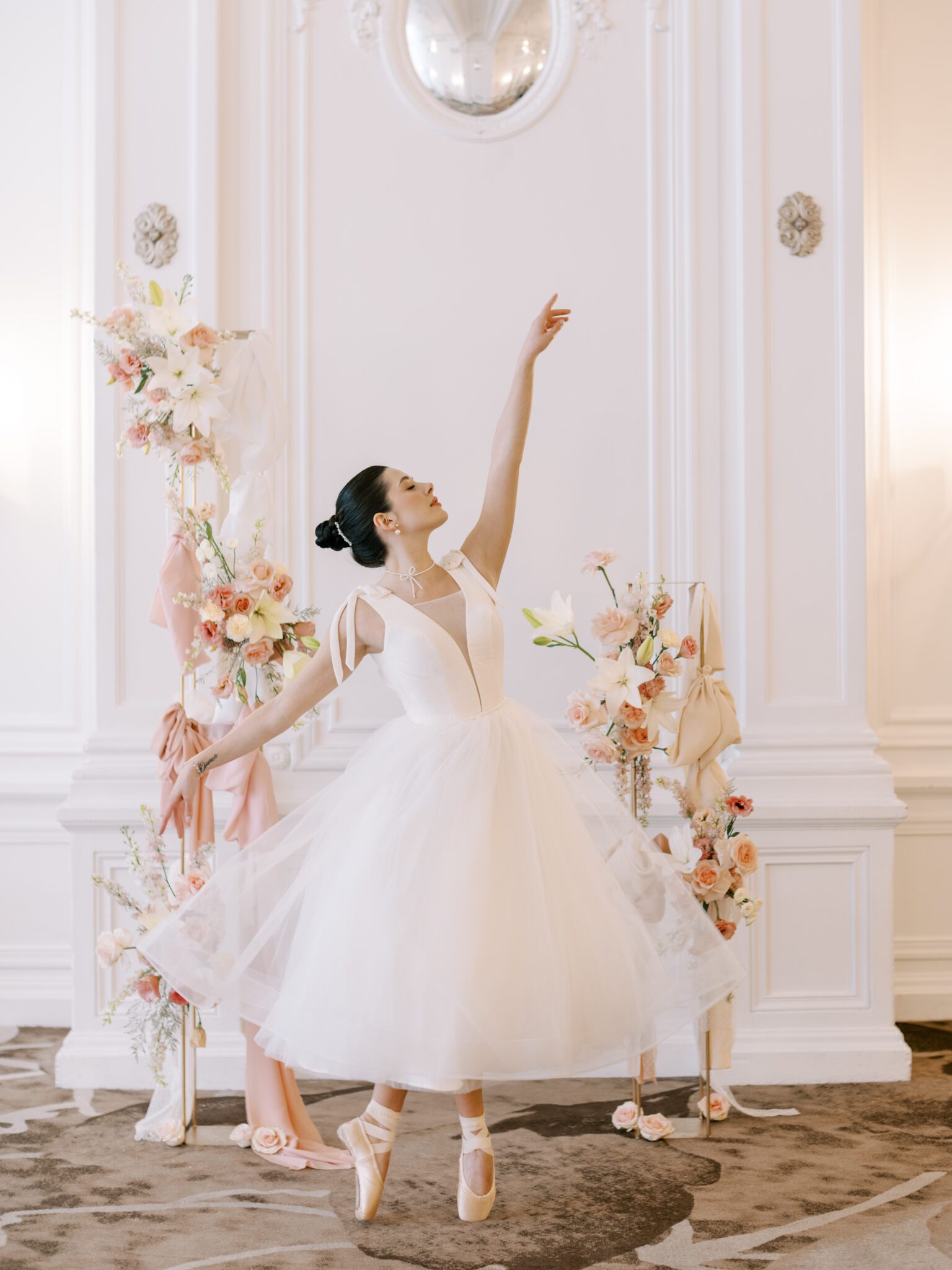 A ballerina bride in a white tutu poses en pointe with arms raised, surrounded by pink and white floral arrangements, in an elegant, ornate room with white walls and a decorative mirror. Whimsical ballet blush pink wedding inspiration at Fairmont Palliser Hotel. 