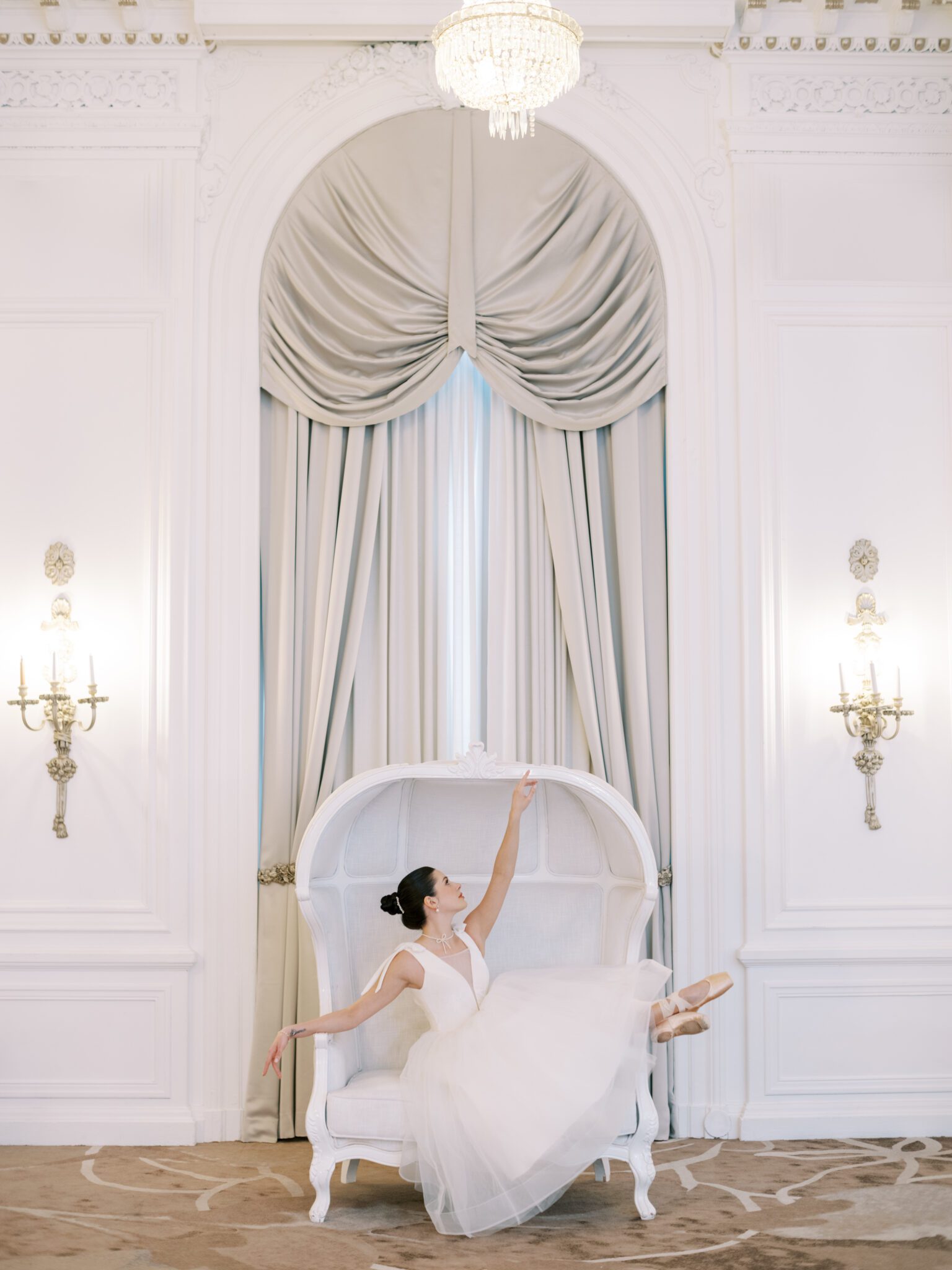 A ballerina in a white tutu sits on an elegant white settee, gracefully adjusting her pink pointe shoe. She has her dark hair in a bun and is surrounded by light-colored, sophisticated decor.
