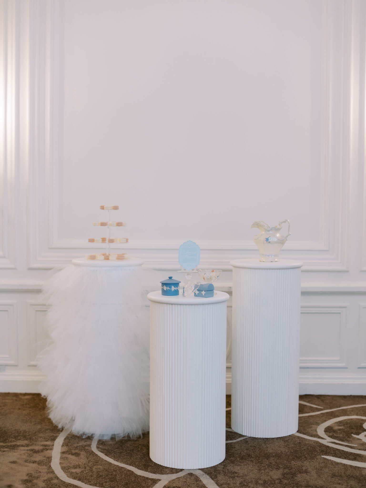 Three white display pedestals hold elegant treats and decor, including blue wedgwood boxes, a tiered tray with handpainted macarons, and vintage decor, in a softly lit room with white paneled walls and a brown carpet.