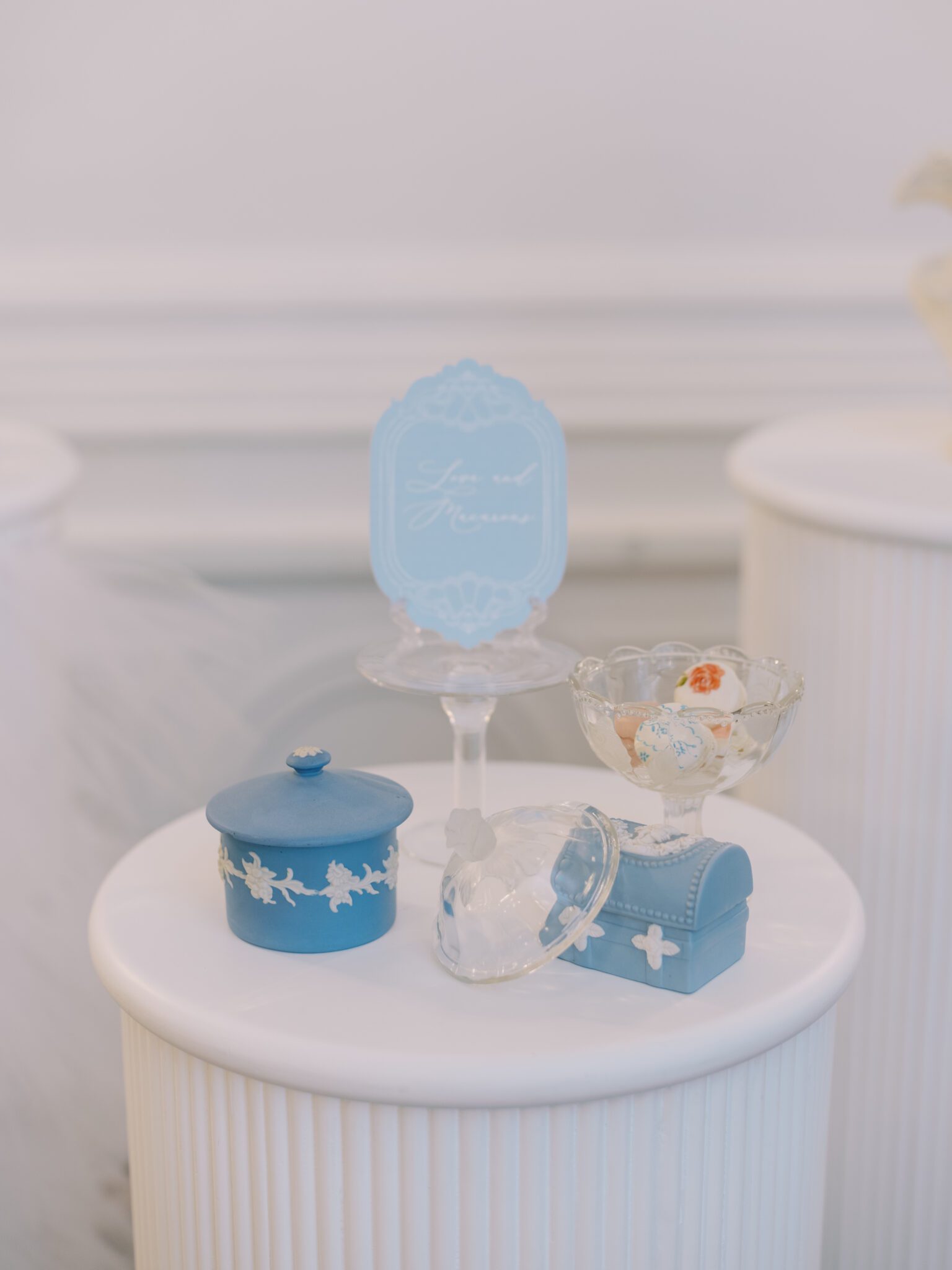 Display pedestals hold elegant treats and decor, including blue wedgwood boxes, a tiered tray with handpainted macarons, and vintage decor, in a softly lit room with white paneled walls and a brown carpet.