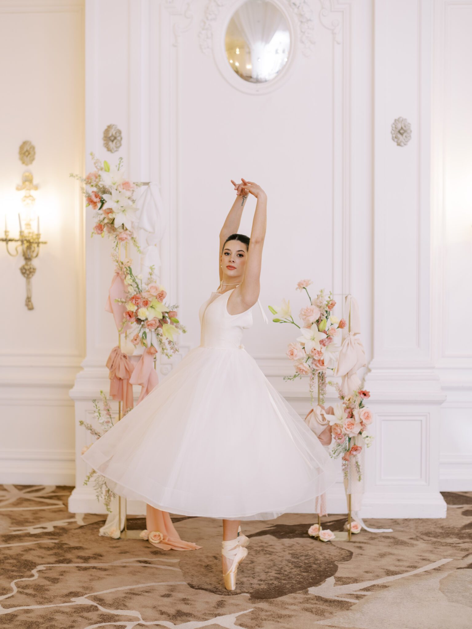 A ballerina bride in a white tutu poses en pointe with arms raised, surrounded by pink and white floral arrangements, in an elegant, ornate room with white walls and a decorative mirror. Whimsical ballet blush wedding inspiration at Fairmont Palliser Hotel. 