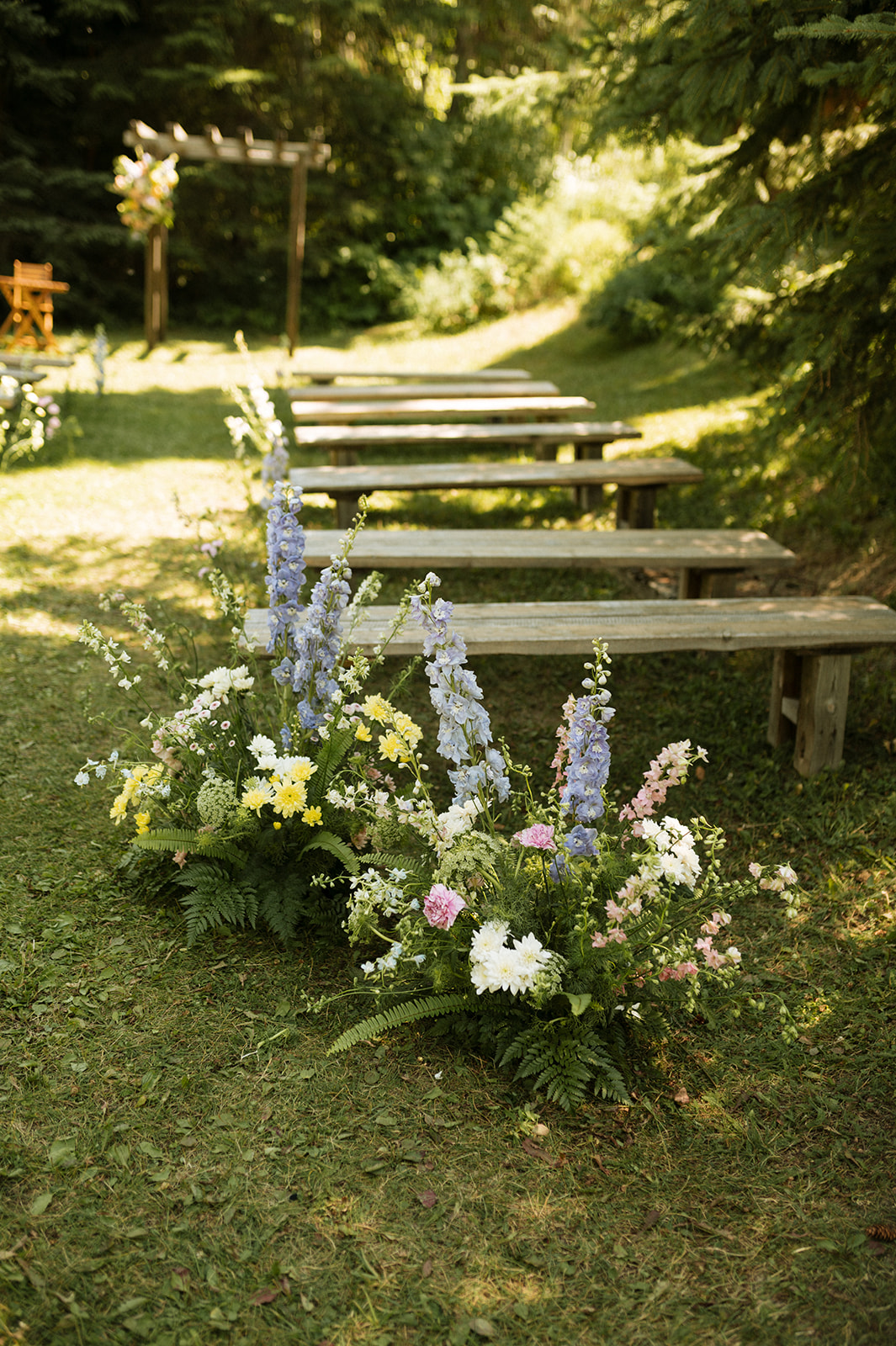 Wooden benches arranged outdoors on grass, lined with lush floral arrangements featuring white, yellow, purple, and pink flowers, with greenery and trees in the background. A wooden arch with flowers is visible in the distance. Wedding Ceremony Inspiration in Golden BC
