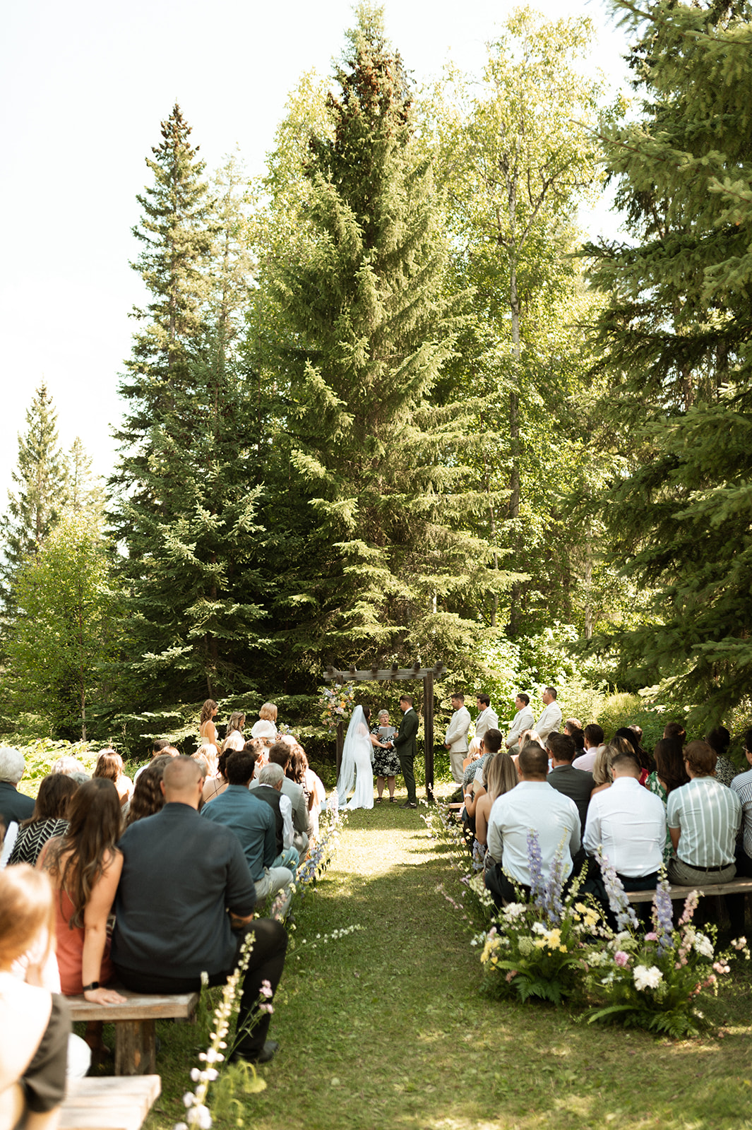 Wooden benches arranged outdoors on grass, lined with lush floral arrangements featuring white, yellow, purple, and pink flowers, with greenery and trees in the background. A wooden arch with flowers is visible in the distance. Wedding Ceremony Inspiration in Golden BC