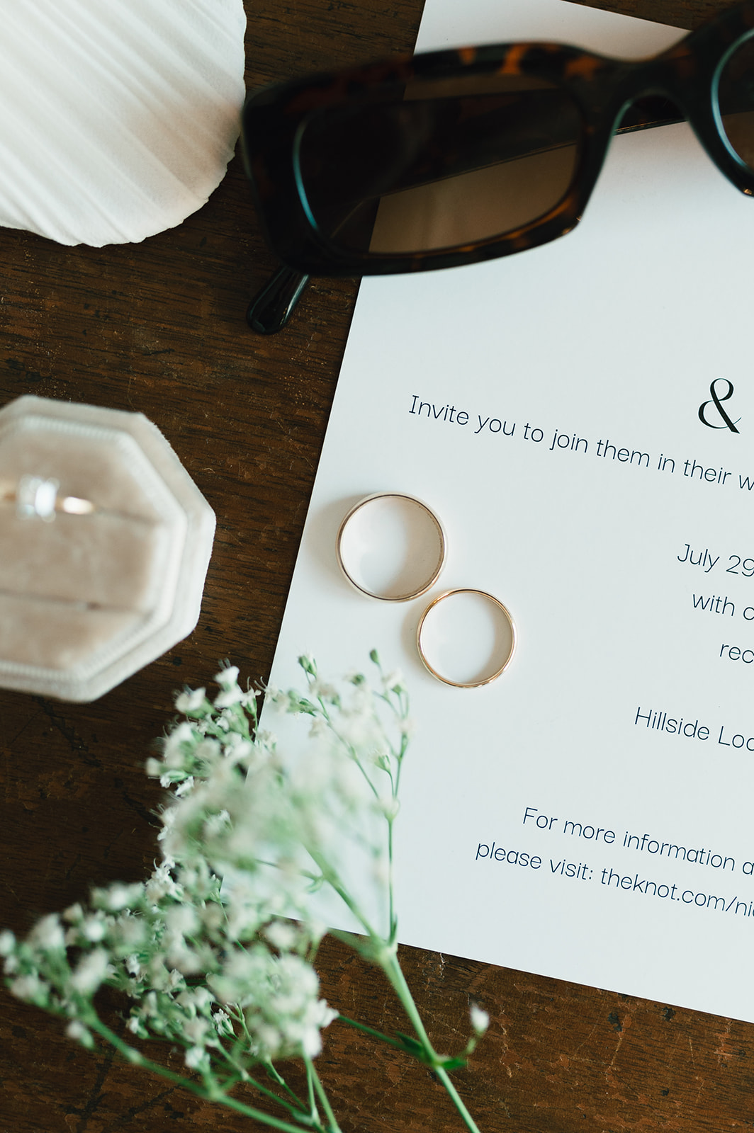 Flat lay of wedding accessories including white bow shoes, sunglasses, wedding invitation, two rings, a ring box with a diamond ring, delicate earrings, and a small sprig of white flowers on a wooden surface.