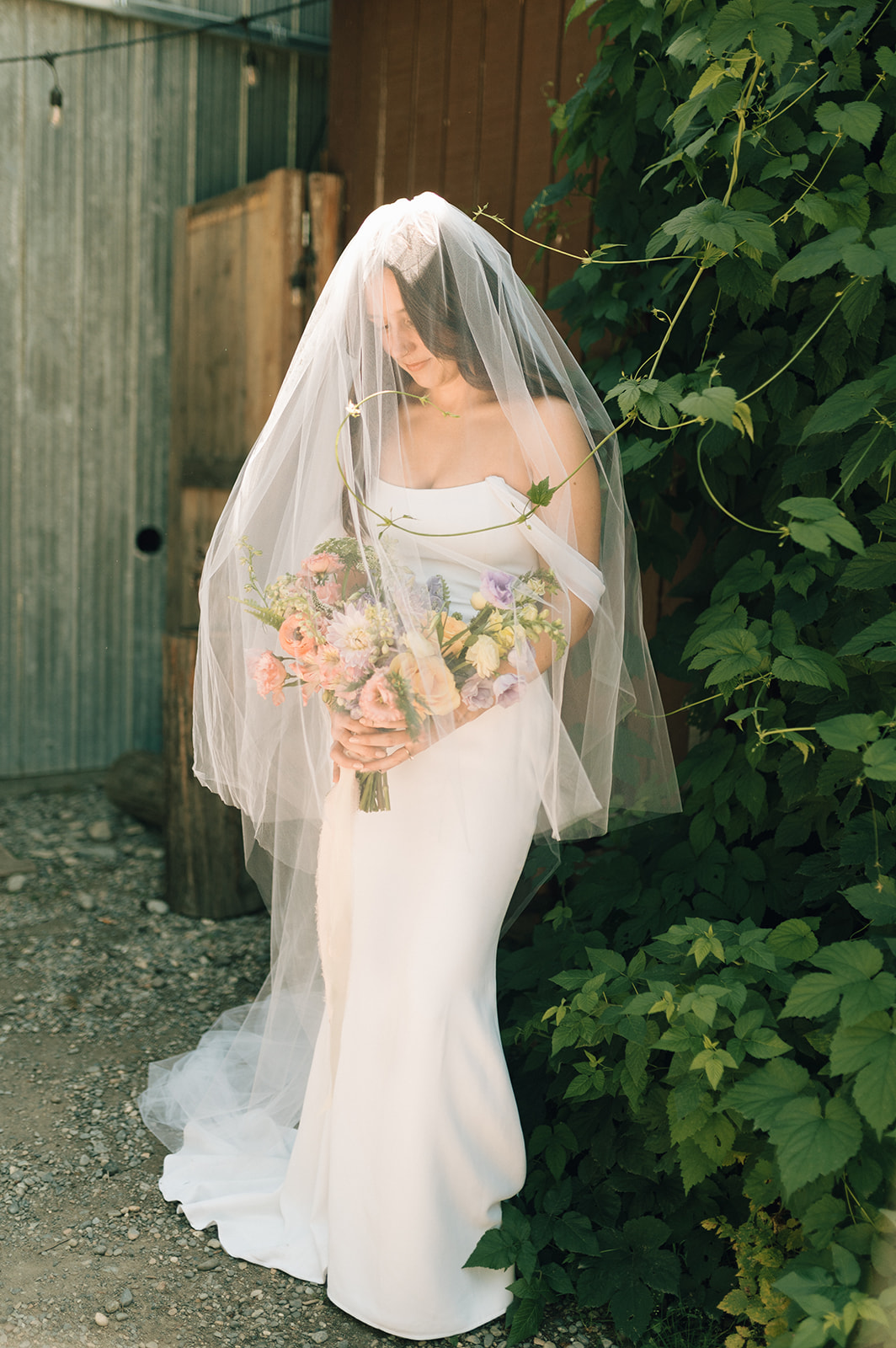 A bride in a white gown and veil stands outdoors, holding a colorful bouquet of flowers. She is surrounded by greenery and sunlight, with rustic wooden and metal structures in the background.