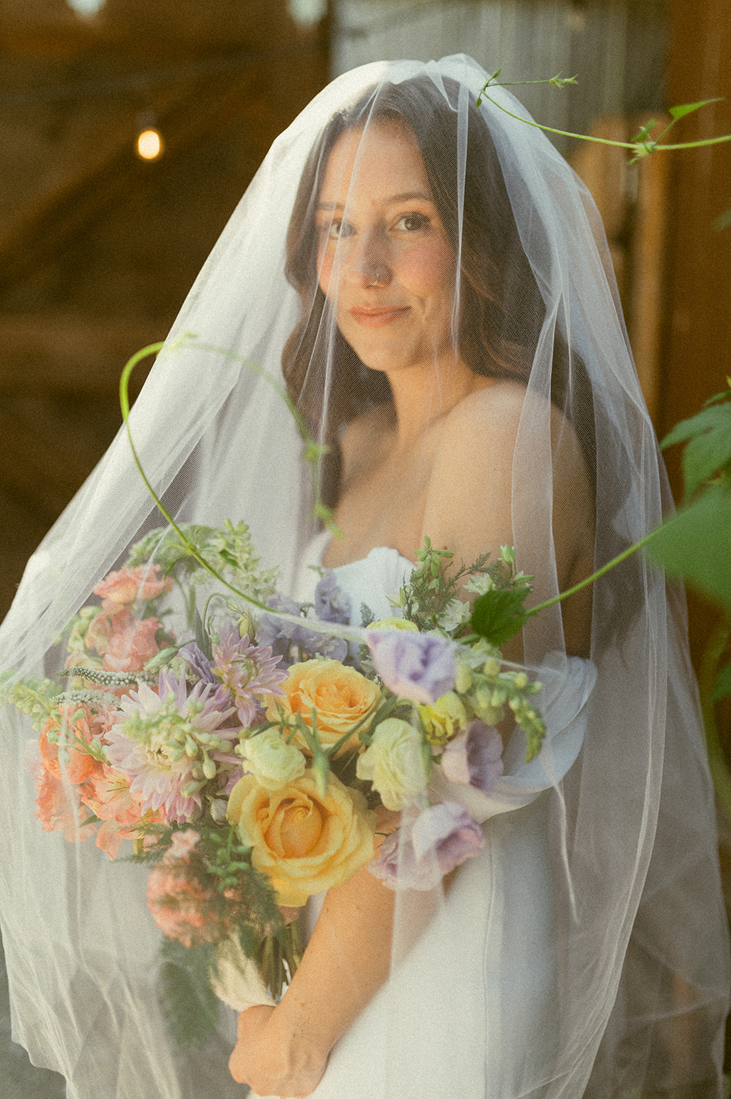 A bride in a white gown and veil stands outdoors, holding a colorful bouquet of flowers. She is surrounded by greenery and sunlight, with rustic wooden and metal structures in the background.