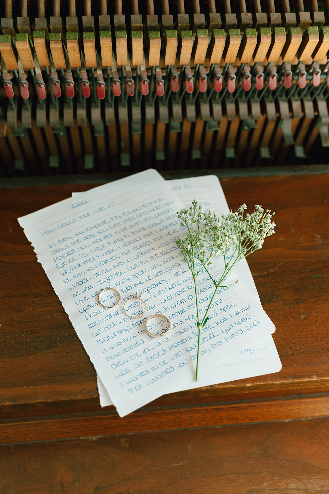 Flat lay of wedding accessories including white bow shoes, sunglasses, wedding invitation, two rings, a ring box with a diamond ring, delicate earrings, and a small sprig of white flowers on a wooden surface.